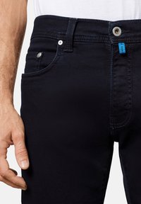 Jeans de denim oscuro con cinco bolsillos, herrajes metálicos y costuras en contraste. Presenta una etiqueta azul en la cinturilla. Textura suave.