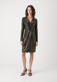 Robe noire à sequins avec un décolleté en V, des manches longues et un design enveloppant. Présente un tissu froncé et une silhouette ajustée. Associée à des escarpins noirs à lacets.