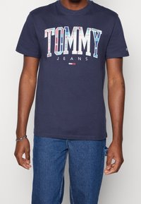 Marinblå bomulls-T-shirt med en stor grafik av "TOMMY JEANS" i flerfärgade bokstäver. Kortsleeves och rundad halsringning, avslappnad passform.
