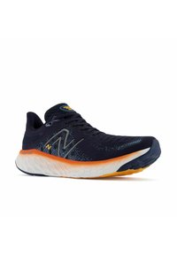 Marineblaue Laufschuhe mit Mesh-Obermaterial, kontrastierender orangefarbener Mittelsohle und strukturiertem weißen Außensohle. Mit dem New Balance-Logo an der Seite.