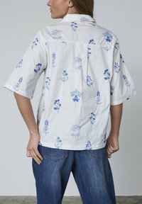 Kortærmet skjorte i hvid bomuld med blå blomsterprint, med krave og rygplissering. Kombineret med blå denim jeans.