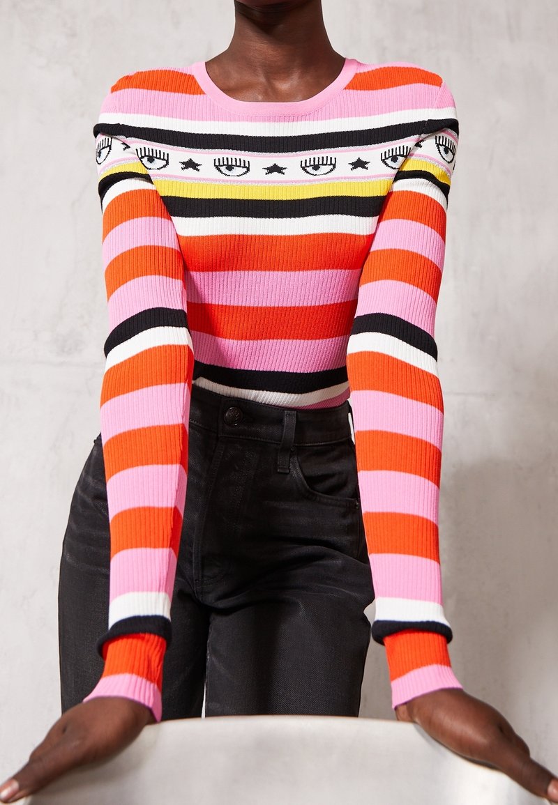 Personne portant un pull à manches longues rayé avec des bandes roses, orange, noires, blanches et jaunes, présentant des motifs d'œil et d'étoile sur la poitrine.