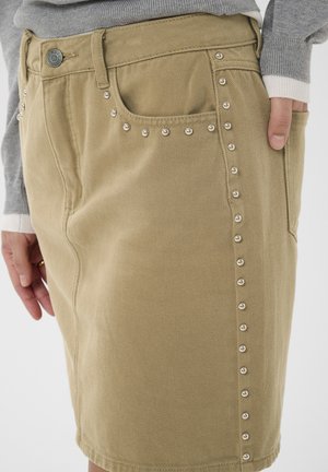 Béžová džínsová sukňa s ozdobnými striebornými cvočkami pozdĺž vreciek a bočného švu, nosená s viditeľnými šedými a bielymi vrstvenými rukávmi.