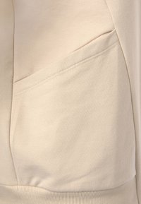 Beigefarbener Sweatshirtstoff mit glatter Textur, der über eine Seitentasche, flache Nähte und gerippte Bündchen am Saum verfügt.