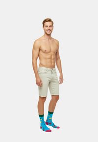 Pantalones cortos de mezclilla beige claro; calcetines coloridos con patrones de frutas en azul, verde y rosa. Sin camiseta, pose de pie, fondo neutral.