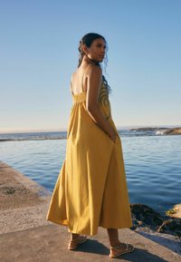 Robe maxi en tissu jaune doux avec un dos élastique, des bretelles fines et une broderie sombre complexe. Présente une coupe en biais et des poches latérales.