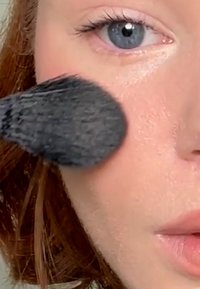 Primo piano di un volto mentre applica il trucco. Il pennello è rotondo e nero, mentre la pelle è chiara e luminosa. Le caratteristiche includono occhi blu e labbra morbide.