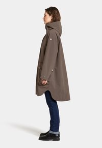 Ruskea vedenpitävä takki hupun kanssa, sivutaskut ja nepparit, päällä tummat farkut ja mustat saappaat, esitelty valkoista taustaa vasten.