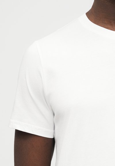 Κοντινό πλάνο ενός ατόμου που φοράει ένα απλό λευκό T-shirt με κοντά μανίκια και στρογγυλή λαιμόκοψη, εμφανίζοντας το πάνω μέρος του κορμού και την περιοχή των ώμων.