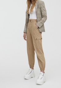 Pantalons cargo beige avec poches latérales, poignets élastiques et tissu lisse, associés à un haut blanc court et un blazer à motifs. Baskets blanches à semelles épaisses.