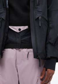 Κοντινό πλάνο ενός ατόμου που φοράει μαύρο μπουφάν πάνω από μαύρο τοπ και ανοιχτό μωβ παντελόνι με τσέπες με φερμουάρ και μαύρη ζώνη με κουμπιά.