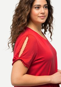 Top corto rojo de manga corta con escote en V y acabado decorativo de pompones en las aberturas de las mangas. Confeccionado en tejido suave y ligero.