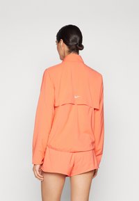 Veste coral Nike avec un col montant et un design à dos superposé. Fabriquée en matériau léger, elle présente un logo blanc au-dessus du milieu du dos.