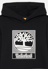 Svart huvtröja med en vit Timberland-logotyp som har ett träd inuti en cirkulär design och en horisontell randig bakgrund.