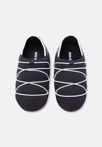 Μαύρα παπούτσια χωρίς κορδόνια με λευκούς ελαστικούς ιμάντες σε σχήμα σταυρού και υφασμένα εσωτερικά πέλματα, απεικονιζόμενα από πάνω σε απλό φόντο.