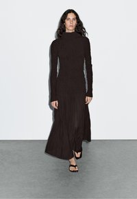 Black long-sleeve dress with a high neck, textured fabric, fitted bodice, and loose skirt. Worn with flat black sandals.
