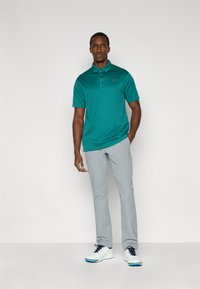 Τιρκουάζ πόλο πουκάμισο με κοντά μανίκια και γιακά· συνδυασμένο με ανοιχτό γκρι παντελόνι και λευκά παπούτσια με μπλε λεπτομέρειες.