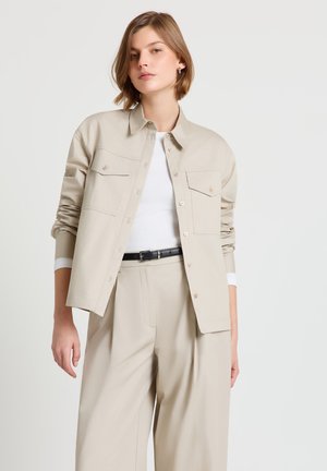 Jeune femme portant une veste beige boutonnée sur un haut blanc et un pantalon large assorti avec une ceinture noire, debout devant un fond uni.