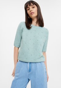 Maglione azzurro chiaro a maniche corte con colletto rotondo, adornato con piccole decorazioni a perline distribuite uniformemente sulla parte anteriore, realizzato in tessuto morbido.