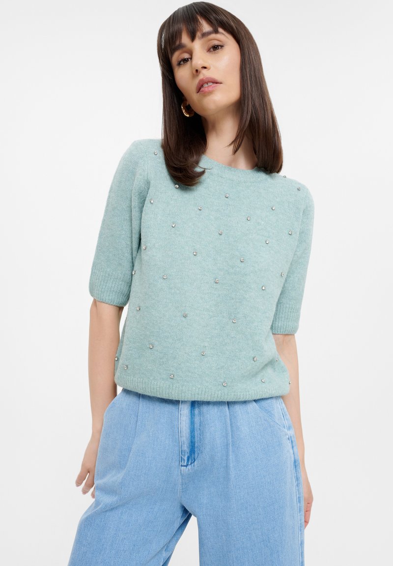 Maglione azzurro chiaro a maniche corte con colletto rotondo, adornato con piccole decorazioni a perline distribuite uniformemente sulla parte anteriore, realizzato in tessuto morbido.