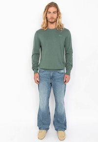 Groen tricot trui met lange mouwen en geribbelde textuur, gecombineerd met lichtblauwe flare jeans en beige mocassins. Simpel, strak ontwerp.