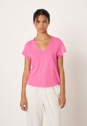 Mlada ženska s kodrastimi lasmi, ki nosi majhne obročaste uhane, roza majico z V-izrezom in svetlo sive hlače, stoji na enobarvnem ozadju.