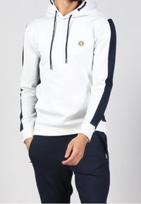Felpa bianca con strisce laterali blu navy, cordini e un logo circolare sul petto. Abbinata a pantaloni blu navy con tasche con zip.