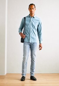 Camisa de botones azul claro, jeans claros ajustados, zapatos negros y un bolso negro de hombro. El modelo está de pie contra un fondo neutro.