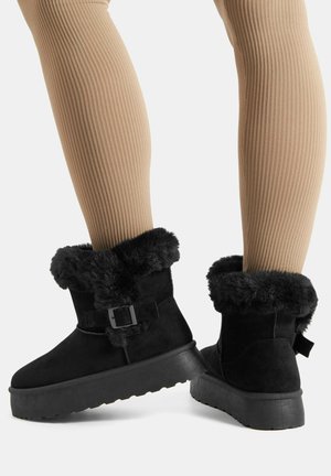 Stivaletti alla caviglia in camoscio nero con bordo spesso in pelliccia, dettaglio con fibbia e suole platform indossati con leggings a coste beige su sfondo uniforme.