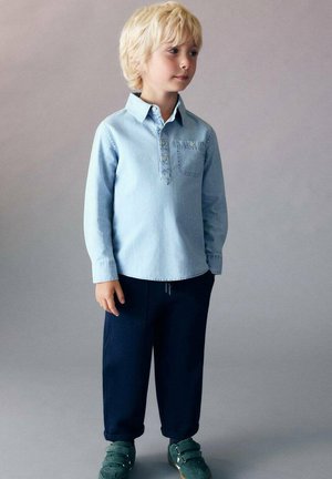Chemise en denim bleu clair à manches longues avec une patte de boutonnage, pantalon bleu marine et chaussures vertes à velcro. Dispose d'une poche poitrine.