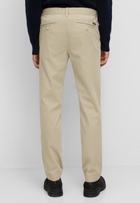 Beige chinos with a straight-leg fit, back pockets with button closures, and a smooth fabric texture, paired with black footwear.