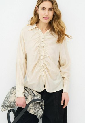 Woman with wavy blonde hair wears a cream ruched blouse and black pants, holding a patterned light jacket with dark straps.