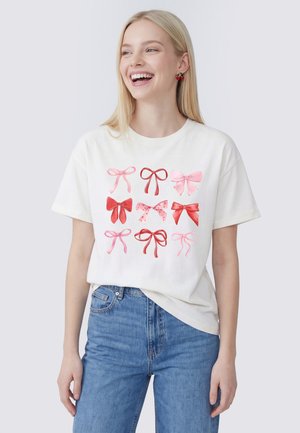 Jonge vrouw met lang blond haar, die een wit t-shirt met negen roze en rode strikjes draagt en een blauwe spijkerbroek, glimlacht en kijkt naar rechts.