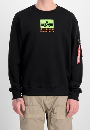 Czarna bluza z neonowo zielonym i pomarańczowym logo "Alpha Industries", beżowe spodnie cargo, czerwona metka "Remove Before Flight" na kieszeni na zamek na lewym rękawie.