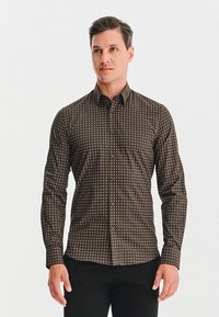 Mężczyzna stoi z luzem na rękach, ubrany w dopasowaną brązową koszulę z małym geometrycznym wzorem i czarne spodnie, zwrócony lekko w prawo.