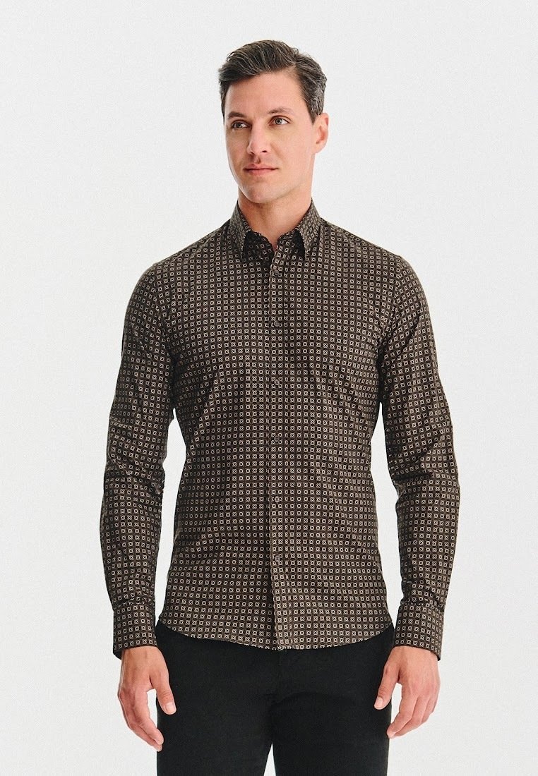 Mężczyzna stoi z luzem na rękach, ubrany w dopasowaną brązową koszulę z małym geometrycznym wzorem i czarne spodnie, zwrócony lekko w prawo.