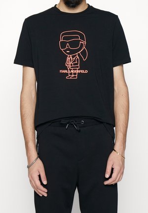Hombre con camiseta negra con figura de dibujo animado en naranja neón de Karl Lagerfeld con gafas de sol y texto "Karl Lagerfeld" en el pecho.