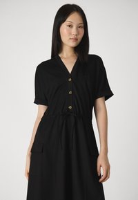 Vestido preto com decote em V, mangas curtas com dobra e cintura com cordão. Apresenta botões dourados e bolsos laterais. Textura de tecido suave.