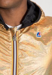 Zlatá kovová lehká bunda s černou kapucí, předním zipem a vícebarevnými pruhovanými detaily. Má nášivku s logem K-Way.