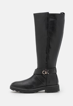 Botte noire en cuir montant jusqu'au genou avec talon bas, bout rond, et une sangle décorative avec un logo "CK" métallique sur le côté extérieur.
