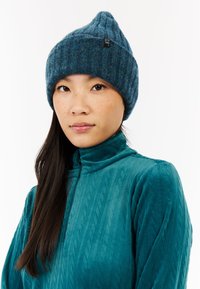 Czapka dzianinowa w ciemnym teal z zaznaczoną górą i subtelnymi wariacjami kolorystycznymi. Noszona z dopasowaną, teksturowaną bluzą z wysokim kołnierzem w odcieniu teal.