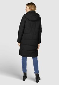 Doudoune noire avec capuche, présentant des coutures horizontales et une longueur plus longue, associée à un jean bleu et des bottines noires.