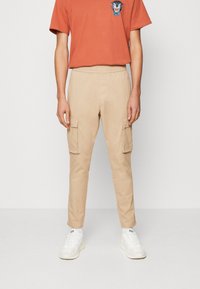 Pantalones cargo beige con piernas ajustadas, dos bolsillos laterales y cinturilla elástica. Combinados con una camiseta naranja con un diseño gráfico.