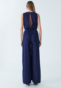 Mornarsko modra kombinezona brez rokavov z elastičnim pasom in odprtim hrbtom. Hlače širokih nog, gladke teksture in minimalističnega dizajna.