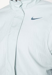 Veste Nike bleu clair avec un col montant, fermeture éclair, et logo Nike swoosh bleu foncé sur la poitrine.