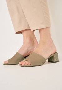 Des mules ouvertes en suede vert avec une large bride et un talon bloc texturé à motif géométrique. Semelle plate, design minimaliste.