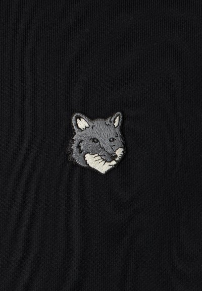 La tela negra presenta una pequeña cabeza de zorro bordada, en tonos grises con acentos en blanco, centrada en el material. Textura suave, diseño minimalista.
