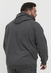 Sudadera gris con una capucha amplia, puños acanalados y cinturilla. Presenta una pequeña etiqueta de marca en la parte inferior izquierda cerca del dobladillo.