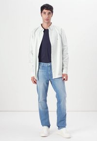 Chemise blanche rayée à boutons sur un t-shirt bleu marine, associée à un jean bleu clair et des baskets blanches. Design simple sans motifs.