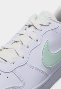 Λευκό αθλητικό παπούτσι με λευκά κορδόνια και λογότυπο Nike swoosh σε ανοιχτό πράσινο στο πλάι, που δείχνει λεπτομερή ραφή και λεία υφή δέρματος.